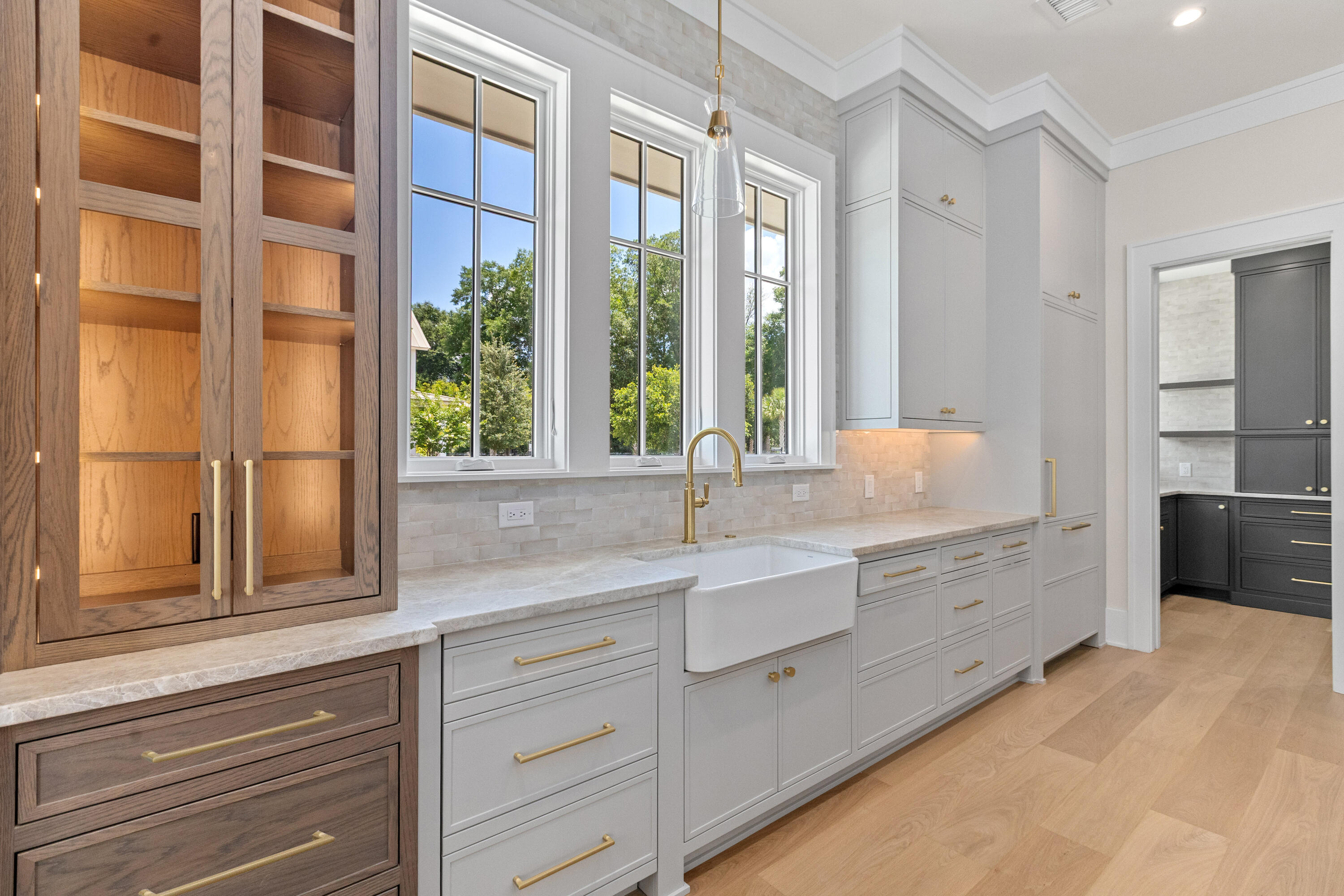 Working scullery with a farmhouse sink under a run of windows, walnut glass-front cabinetry, grey lower cabinets, and a darker pantry beyond