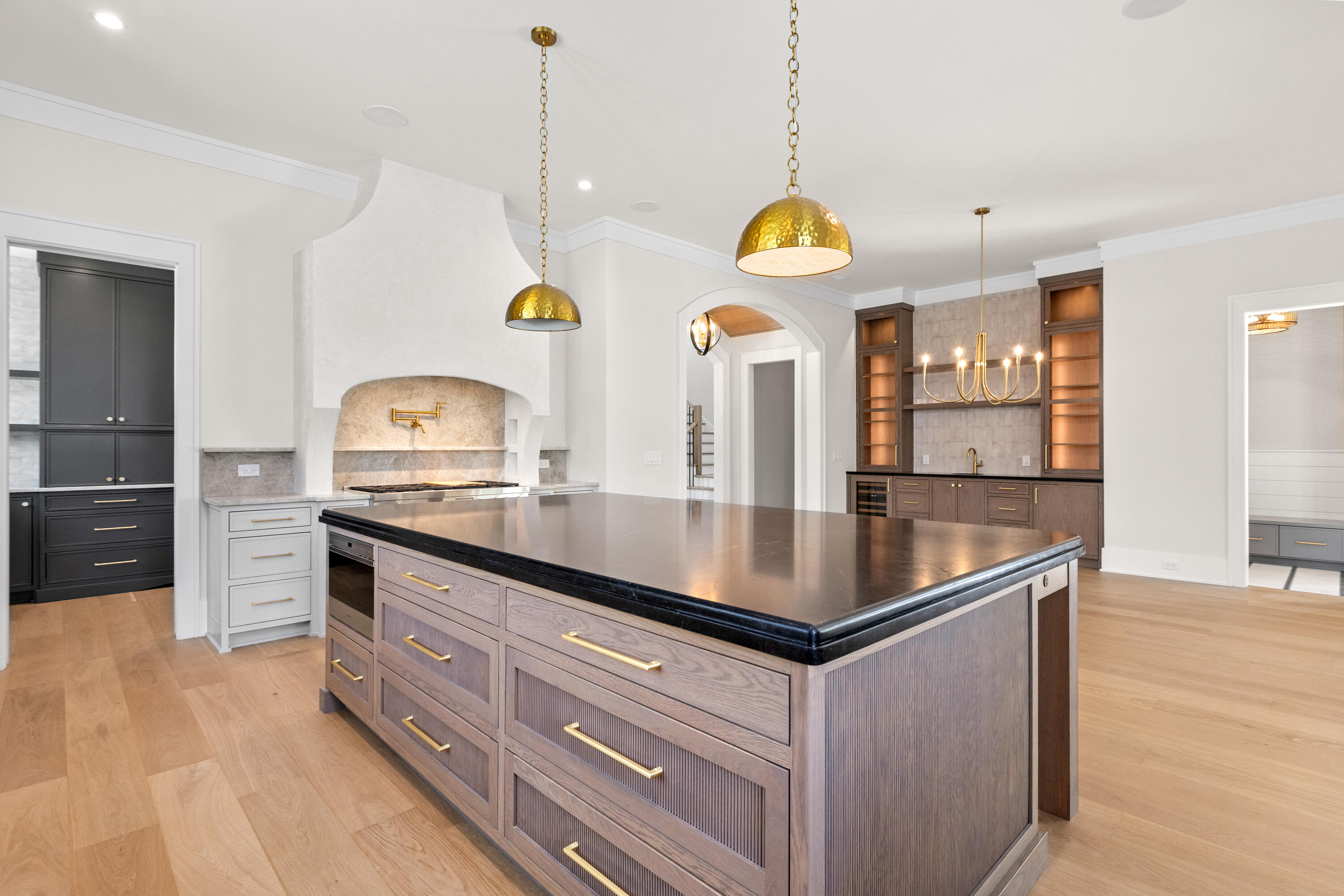 Open kitchen with a large walnut island topped in honed black stone, two hammered-brass dome pendants overhead, and a bar wall beyond
